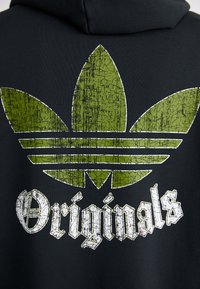 Svart huvtröja med ett slitigt grönt trebladigt logotyp och vit text "Originals", med en sprucken textur och djärv design.