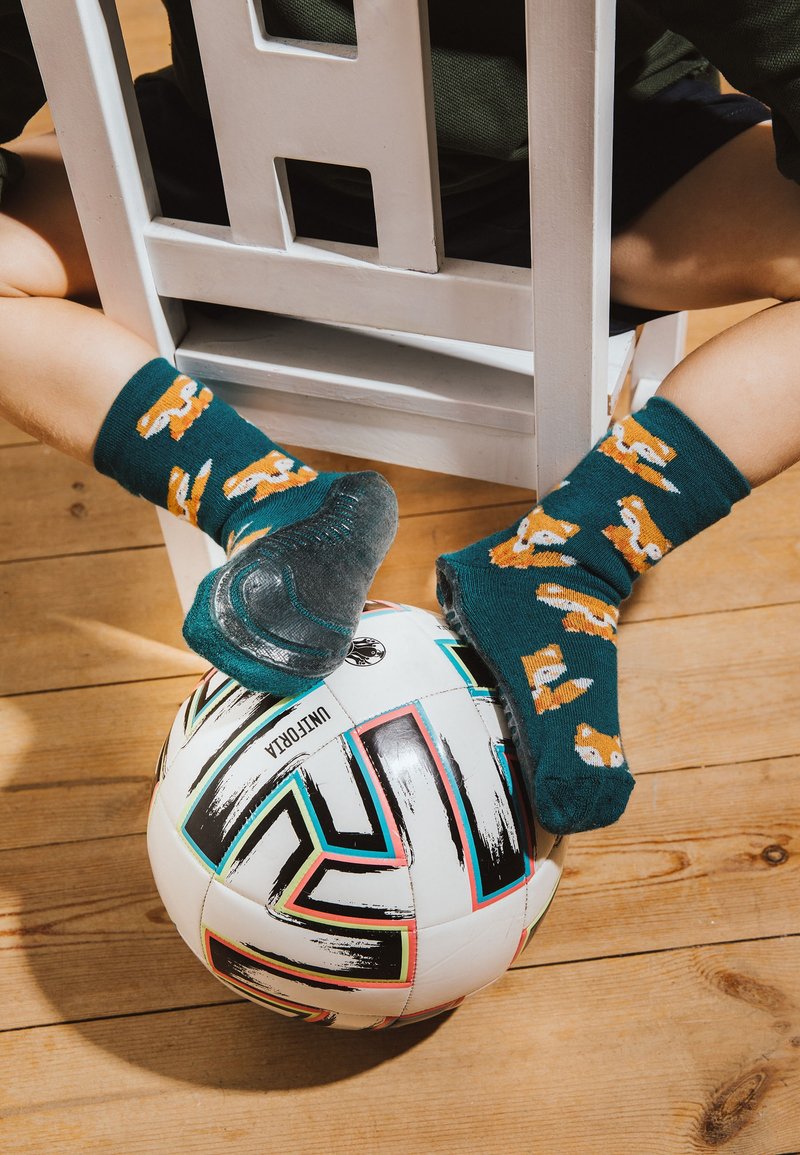 Kind mit türkisfarbenen Socken mit Fuchsmustern sitzt hinter einem weißen Stuhl, die Füße ruhen auf einem schwarz-weißen Fußball auf einem Holzboden.