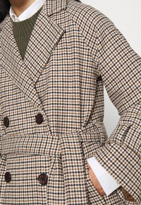 Dubbelzijdige jas in een bruine en crèmekleurige houndstoothpatroon, met grote bruine knopen, een gestrikt riem en brede revers.