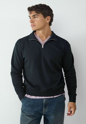 Jeune homme aux cheveux bouclés portant un sweat-shirt noir à fermeture éclair jusqu'au quart, une chemise violet clair en dessous, et un jean bleu, regardant vers la gauche.