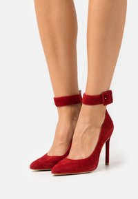 Escarpins en daim rouge avec un bout pointu, une bride de cheville et un talon aiguille. Texture lisse avec une couleur vive.
