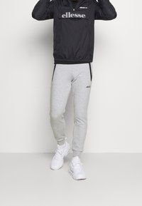 Sort vindjakke med lynlås, grå sweatpants og hvide sneakers. Vindjakken har et logo og elastiske manchetter; sweatpantsene har sidelommer.