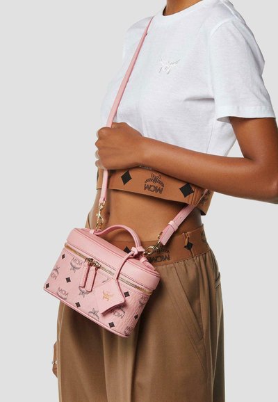 Femme portant un crop top blanc et un pantalon marron, tenant un sac bandoulière rose à motifs MCM avec des fermetures éclair dorées et une poignée supérieure.