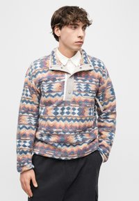 Νεαρός άνδρας που φοράει πολύχρωμο φλις πουλόβερ με σχέδια, με ψηλό γιακά και κουμπιά πατιλέτας, συνδυασμένο με σκούρο παντελόνι, κοιτώντας προς τα πλάγια.