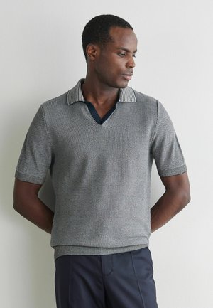 Mann trägt ein kurzärmliges, strukturiertes graues Strick-Poloshirt mit dunklem Kragen, steht vor einer hellen Wand, schaut zur Seite, die Hände hinter dem Rücken verschränkt.