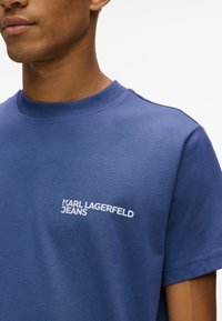 Zils kokvilnas T-krekls ar apaļu izgriezumu, uz kura kreisajā krūtī ir balts "KARL LAGERFELD JEANS" logo, īsām piedurknēm un gludu tekstūru.
