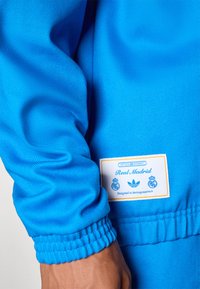 Φωτεινό μπλε αθλητικό μπουφάν με ριμπ μανσέτες, με ετικέτα που γράφει «Real Madrid» και λεπτομέρειες επωνυμίας. Ομαλή υφή υφάσματος.