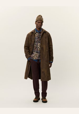 Brown leather long coat, floral patterned shirt, blue denim collared shirt, burgundy pants, brown boots, and a beige knit beanie.