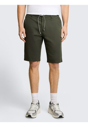 Mann trägt olivgrüne knielange Shorts mit Kordelzug, weiße Socken und weiße Turnschuhe, steht vor einem einfarbigen Hintergrund.