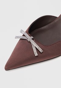 Chaussure plate marron à bout pointu avec un nœud décoratif en strass argenté sur le dessus, près de la zone des orteils.