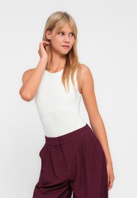 Body bianco senza maniche con scollo alto, abbinato a pantaloni alti di un profondo borgogna con pieghe e tasche laterali.
