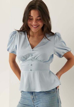 Vrouw glimlacht naar beneden kijkend, draagt een lichtblauw blouse met pofmouwen, knopen en peplumzoom, gecombineerd met lichtblauwe spijkerbroek, handen in de achterzakken.