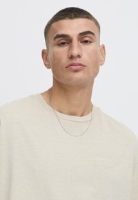 Cream-colored cotton t-shirt with a round neckline, featuring a subtle embroidered logo. A silver chain necklace adds a metallic accent.