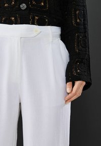 Schwarze gehäkelte Strickjacke mit runden Mustern, kombiniert mit weißen, plissierten Hosen mit Knopfverschluss und glatter Textur.