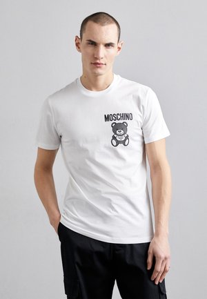 Camiseta de algodón blanca con un gráfico negro de un oso de peluche y el texto "MOSCHINO". Mangas cortas, corte estándar, textura suave.