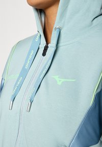 Lys blå hettegenser med grønn detaljer. Har snor i hetten, Mizuno-logo og kontrasterende paneldesign. Glatt stoffoverflate.