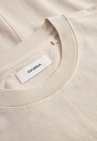 Beige Baumwoll-T-Shirt mit Rundhalsausschnitt. Präsentiert ein Etikett mit der Aufschrift "GABBA". Glatte Textur mit einem schlichten Design und gerippten Kanten.