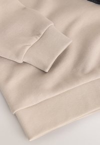 Manche et ourlet inférieur de sweat-shirt beige avec poignets côtelés, tissu doux texturé et légères plis visibles sur le matériau.