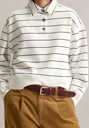 Polo blanco de manga larga con finas rayas horizontales negras y tres botones marrones, llevado con pantalones marrones y un cinturón marrón oscuro.