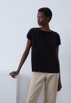 T-shirt noir plissé à manches courtes et col rond, associé à un pantalon beige. Texture lisse avec un détail de drapé subtil.