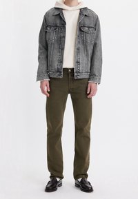 Chaqueta vaquera en negro desgastado con dos bolsillos en el pecho, combinada con una sudadera ligera con capucha. Pantalones verde oliva y zapatos negros completan el conjunto.