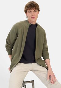 Olivengrøn zip-up cardigan med ribbet tekstur og stående krave, lagt over en sort skjorte, parret med lysebeige bukser.