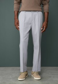 Light gray tailored trousers with front pleats, smooth texture, and a straight cut. Paired with beige suede sneakers.