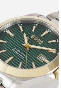 Cadran de montre à rayures vertes avec des accents en or, bracelet en acier inoxydable argenté, étanche jusqu'à 50 mètres, avec une fenêtre de date à 6 heures.