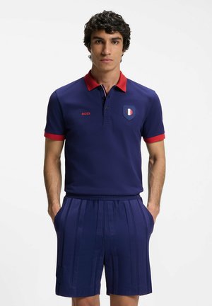 Uomo in piedi con le mani nelle tasche, indossa una polo blu navy con colletto e polsini rossi e pantaloncini blu navy abbinati.