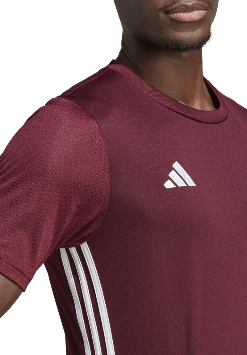Camiseta deportiva burdeos de manga corta. Presenta un diseño de tres rayas blancas en los lados y un logo blanco en el pecho. Tejido texturizado.