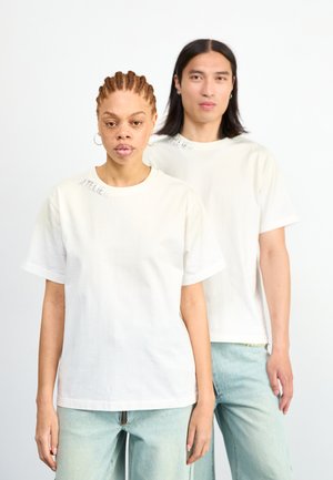 Deux individus portent des t-shirts en coton de couleur écrue, à coupe décontractée et à manches courtes. Un des t-shirts affiche l'inscription "ATELIER" imprimée sur l'épaule.