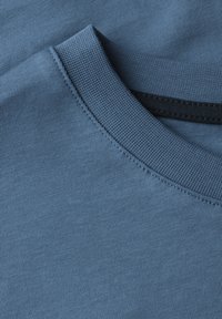 Modrá bavlněná tričko s kulatým výstřihem, žebrovaným límcem a hladkým povrchem. Přišité detaily jsou patrné podél límců a okrajů.