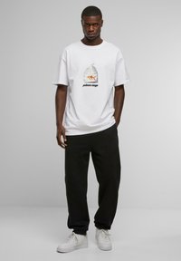 Hvid t-shirt med et trykt grafik af en fisk i en pose med teksten "poisson rouge" nedenfor. Bæres med sorte bukser og hvide sko.