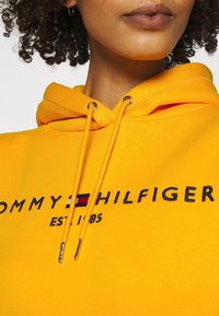 Gelbes Kapuzensweatshirt mit einem Frontlogo, das "TOMMY HILFIGER" und "EST 1985" in Marineblau sowie gestickten Akzenten zeigt. Metallische Enden an den Kordeln.