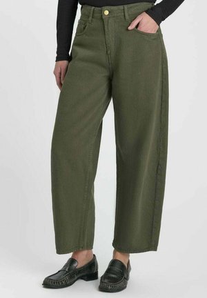 Donna con pantaloni larghi verde oliva, mocassini neri e top nero a maniche lunghe, una mano in tasca su sfondo semplice.