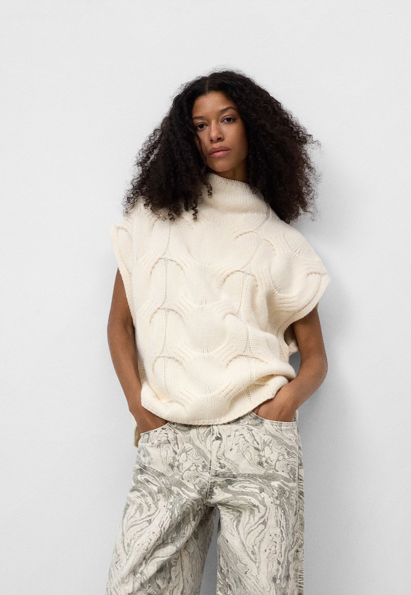 Kremowy sweter z dzianiny z wysokim kołnierzem i teksturowanym wzorem falistym, w połączeniu z jasnymi, marmurkowymi spodniami o szerokich nogawkach.
