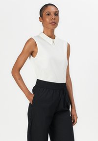 Blusa bianca senza maniche con colletto classico, abbinata a pantaloni neri con vita elasticizzata e tasche laterali. Tessuto a trama liscia.