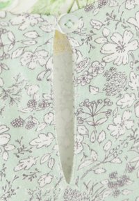 Gros plan sur un tissu floral vert clair avec des fleurs blanches et grises et un petit bouton blanc fermant une encolure en forme de trou de serrure.