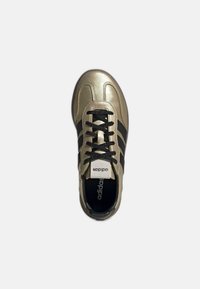 Zapatilla deportiva dorada con rayas negras, punta redonda y cordones negros. Parte superior texturada y lengua con logotipo. Vista superior.