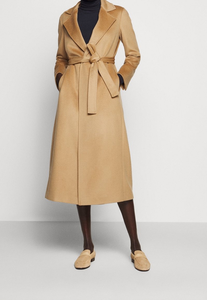 Long manteau en laine beige avec une ceinture nouée porté sur un top noir, associé à des collants foncés et des chaussures beiges sans lacets.
