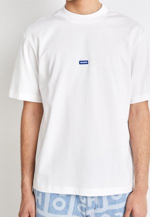 Person iført en ensfarvet hvid kortærmet T-shirt med lille blåt "HUGO" logo på brystet og mønstrede blå og hvide bukser.