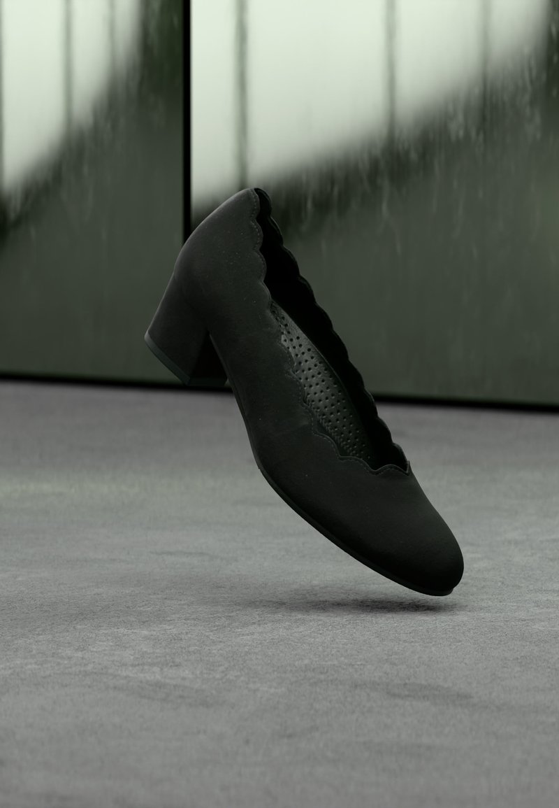 Chaussure femme en daim noir à talon moyen, ornée de bords festonnés et d’une semelle intérieure perforée, présentée sur un fond gris et vert atténué.