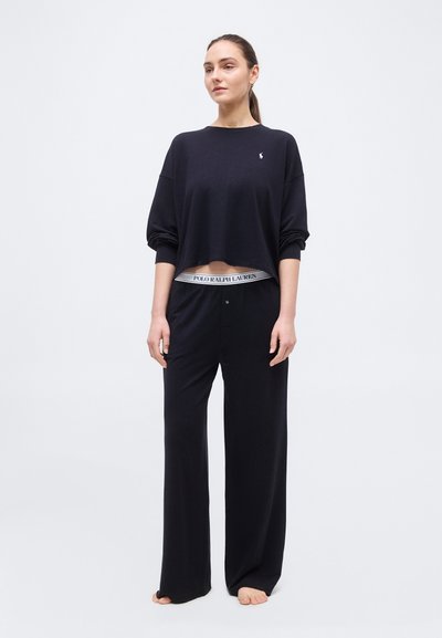 Γυναίκα που στέκεται ξυπόλητη φορώντας μαύρο μακρυμάνικο κοντό τοπ και παντελόνι με φαρδιά πόδια, με ορατή μέση με λογότυπο Polo Ralph Lauren.