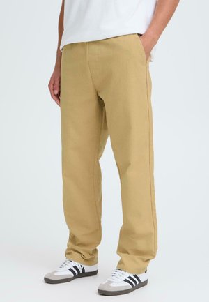 Beige cotton trousers with a straight cut, two side pockets, and subtle stitching details, paired with white and gray athletic shoes.
