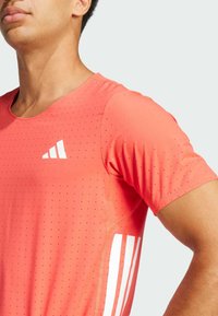 Camiseta deportiva de manga corta en un brillante color coral con textura perforada, que presenta un logo blanco y rayas en los laterales para ventilación y detalle de diseño.