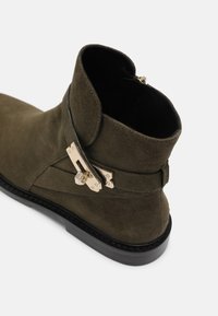 Bottine en suede vert olive foncé avec semelle noire et détail de boucle métallique dorée sur le côté, sur fond blanc.