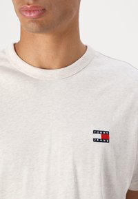 Light gray cotton t-shirt with a round neckline. Features a small blue, red, and white logo patch reading "TOMMY JEANS" on the left chest.