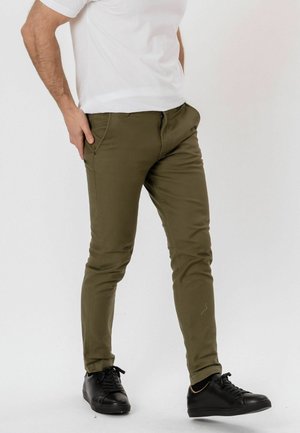Man draagt olijfgroene slim-fit broek, wit t-shirt en zwarte sneakers, met één hand in de zak, staand tegen een effen achtergrond.