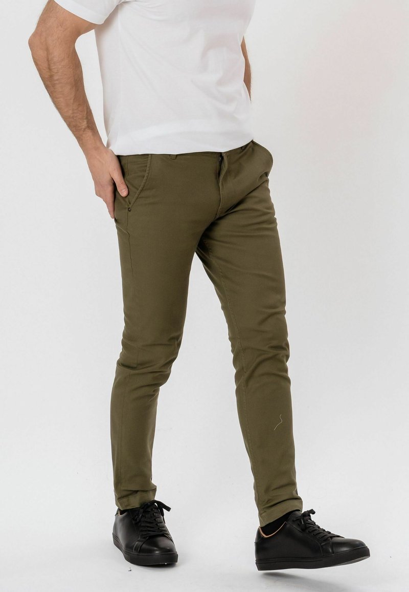 Homme portant un pantalon slim vert olive, un t-shirt blanc et des baskets noires, une main dans la poche, debout devant un fond uni.