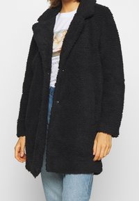 Abrigo negro y peludo confeccionado con una tela texturizada, que presenta un cuello con solapa en muesca, apertura frontal y mangas largas, combinado con una camiseta gráfica debajo.